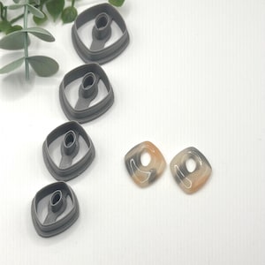 May include: A collection of gray polymer clay cutters and two square earrings. The cutters are square with a rounded interior shape. The earrings are square with a hole in the center, and have a marbled pattern of gray, tan, and white.