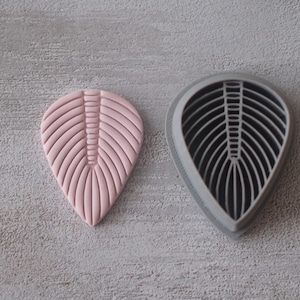 Peut inclure: Un objet rose en forme de feuille avec un motif nervuré à côté d'un emporte-pièce gris en forme de feuille. Les objets sont sur une surface texturée, gris clair. Le design ressemble à un fossile ou à une feuille stylisée.