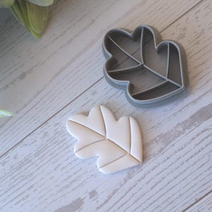 May include: A leaf-shaped cookie cutter and a leaf-shaped cookie. The cookie cutter is gray, and the cookie is off-white with subtle lines. The items are on a light wood-grain surface, with greenery in the background.