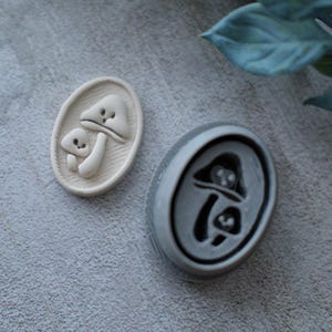 May include: Two mushroom-shaped stamps. One is a light beige color with a raised design of two mushrooms inside an oval. The other stamp is gray with a recessed mushroom design. The stamps are on a gray surface.