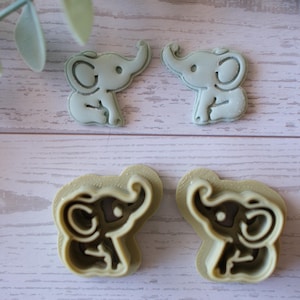 Puede incluir: Cuatro cortadores de galletas con forma de elefante. Dos son de color azul claro y dos de color beige. El diseño del elefante presenta orejas grandes, una trompa y una cara sonriente. Los cortadores están sobre una superficie de vetas de madera clara.