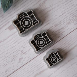 May include: Three grey plastic camera-shaped cookie cutters of varying sizes are arranged diagonally on a light wood-grain surface. The cutters have a detailed camera outline with a lens and buttons.