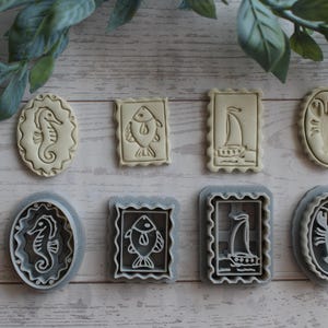 May include: A set of four nautical-themed cookie cutters and their corresponding stamps. The shapes include a seahorse, a fish, a sailboat, and a lobster. The stamps are off-white, while the outer cutter frames are a light gray plastic material.