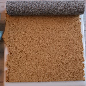 May include: A textured, tan-colored material is partially rolled with a gray roller on a white surface. A sprig of green leaves is in the background. The material has a rough, porous texture.