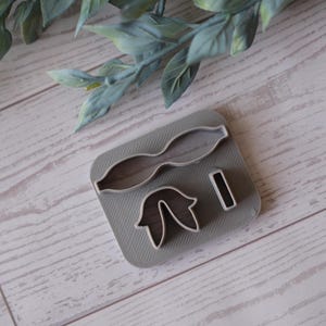 May include: A gray polymer clay bow-shaped cutter with two other shapes, likely for crafting. The cutter is on a gray base, with a wooden background and green leaves in the background.