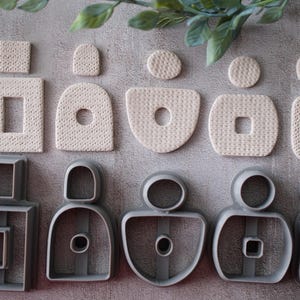 May include: An overhead shot displays a collection of beige clay shapes and matching gray metal cookie cutters. The clay pieces include squares, rectangles, circles, and abstract forms. The cutters mirror the shapes, creating a cohesive set for crafting.