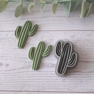 Puede incluir: Tres artículos con forma de cactus sobre una superficie de madera clara. Dos son verdes, texturizados y tridimensionales. El tercero es un contorno en blanco y negro, posiblemente un cortador de galletas o un sello. Hojas verdes en el fondo.