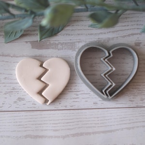 Könnte beinhalten: Ein Ausstecher für ein gebrochenes Herz und ein herzförmiger Keks. Der Ausstecher ist grau und der Keks ist cremefarben. Die Herzform ist in der Mitte mit einem Zickzackmuster geteilt. Die Artikel befinden sich auf einer hellen Holzmaserung.
