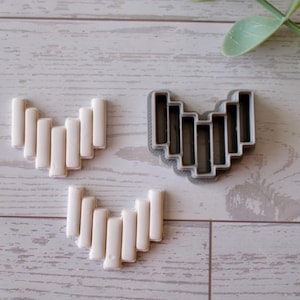 May include: A gray plastic cookie cutter in the shape of a heart with vertical bars. Three sets of white, rectangular, baked goods are arranged in the shape of a heart. The items are on a light wood grain surface.
