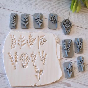 May include: A collection of gray rectangular clay stamps featuring various botanical designs, including leaves, flowers, and a lightbulb shape. The stamps are arranged near a piece of clay with matching embossed floral patterns.