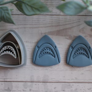 Puede incluir: Un cortador de galletas gris claro con forma de tiburón y dos recortes con forma de tiburón hechos de arcilla azul. El cortador presenta el contorno detallado de una cabeza de tiburón con dientes afilados, y las dos piezas de arcilla azul replican este diseño.