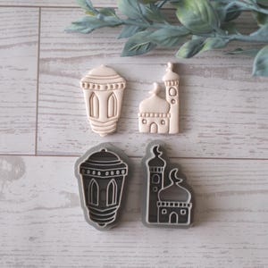 Può includere: Quattro formine per biscotti a forma di lanterna e moschea. Due sono beige e due grigie con interni bianchi. Le formine sono disposte su una superficie chiara con venature del legno e fogliame verde sullo sfondo.