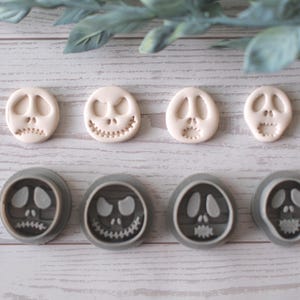 Puede incluir: Una vista aérea muestra ocho cortadores de galletas con el tema de Jack Skellington. Cuatro son blanquecinos y cuatro grises. Los cortadores están dispuestos en dos filas de cuatro, cada uno con una expresión facial diferente. El fondo es de madera clara.