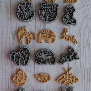 May include: A selection of cookie cutters in animal shapes, such as birds, foxes, rabbits, horses, and an elephant. The cutters are in tan and grey. The designs feature detailed folk art patterns.