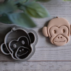 May include: A monkey-shaped cookie cutter and a baked cookie. The cookie cutter is gray, and the cookie is light brown. The monkey has a smiling face, round ears, and a small nose. The cookie cutter is made of plastic.