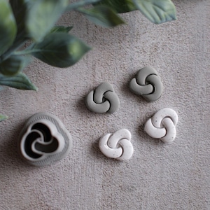 May include: A collection of gray and white clay knot-shaped beads and a matching cutter. The beads are in various shades of gray and white. The cutter is a light gray color with a black interior.