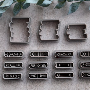 以下が含まれることがあります： さまざまな形の灰色のプラスチック製クッキーカッターのコレクション。カッターには、長方形と装飾的なデザインの長方形が含まれます。デザインには、幾何学模様、ドット、線が含まれます。緑の葉の小枝が背景にあります。
