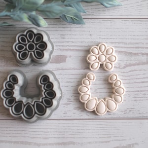 May include: Four floral-shaped cookie cutters. Two are grey with black details, and two are beige. The cutters are arranged on a light wood-grain surface, with a sprig of green leaves in the background. These are ideal for baking.