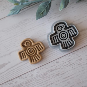 Puede incluir: Dos cortadores de galletas con forma de pájaro. Uno es de color marrón claro, el otro gris. Ambos tienen un diseño similar con una silueta de pájaro, con un ojo circular y detalles de alas. Los cortadores están sobre una superficie de vetas de madera clara.