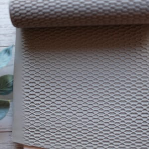 May include: A gray clay roller with a textured honeycomb pattern is shown partially rolled over a sheet of clay. The clay sheet also has the same honeycomb pattern. A sprig of green leaves is in the background.