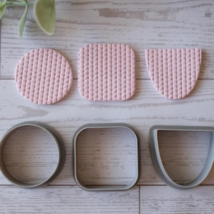 May include: A collection of three pink textured clay shapes: a circle, a square, and a half-circle. Below are three silver metal cookie cutters in matching shapes. The items are arranged on a light wood-grain surface.