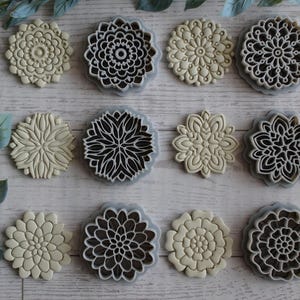 May include: A collection of twelve circular and floral-shaped cookie cutters arranged in three rows. Each cutter features a distinct geometric or petal-like pattern in a light beige color with grey edges, designed for creating intricate impressions.
