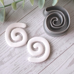 May include: Two white, spiral-shaped objects and a grey spiral-shaped cookie cutter are arranged on a light wood-grain surface. A sprig of green leaves is also present. The cookie cutter is made of plastic.