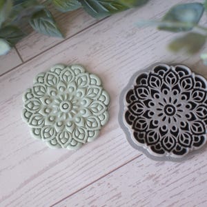 Puede incluir: Dos cortadores de galletas con forma de mandala. Uno es de color verde claro y el otro es blanco y negro. Los cortadores tienen un diseño floral detallado. Los cortadores están sobre una superficie de madera clara.