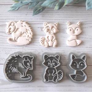 Puede incluir: Un surtido de cortadores de galletas con forma de animales, incluyendo un erizo, un mapache y un zorro. Los cortadores están en dos estilos: uno con un diseño de relieve detallado y otro con un contorno simple. Los colores son beige claro y gris.