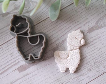 May include: A gray plastic llama-shaped cookie cutter next to a white llama-shaped cookie. The cookie cutter has a detailed outline of the llama's body, including its head, legs, and saddle. The cookie has a textured surface.