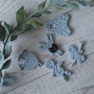 May include: A collection of light blue decorative figurines, including a bunny with a black bow, a voodoo doll, a palette, and a figure with a nail. The figurines are arranged on a light wood surface with green leafy branches.