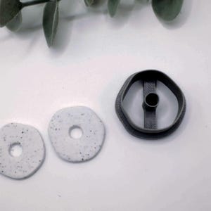 May include: A dark gray cookie cutter and two light gray, speckled, circular clay pieces with holes. The cookie cutter is next to the clay pieces. The items are on a white surface.