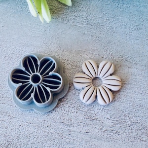 Puede incluir: Un cortador de galletas gris con forma de flor junto a una manualidad terminada con forma de flor. El cortador tiene un diseño detallado de pétalos. La manualidad terminada es blanquecina con detalles marrones.