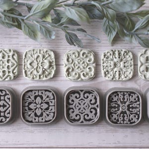 Op de afbeelding: Tien vierkante, decoratieve stempels met bloemen- en geometrische patronen. De bovenste rij heeft lichtgroene stempels, terwijl de onderste rij donkergrijze stempels heeft met een grijze rand. De stempels zijn gerangschikt op een licht houten oppervlak met groene bladeren.