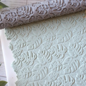May include: A gray rolling pin with a leaf pattern is shown over a light green clay sheet, also imprinted with the leaf design. The clay is on a white surface, suggesting a craft project.