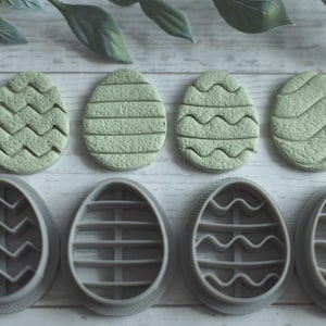 May include: Four green Easter egg-shaped cookies with textured designs sit above four gray egg-shaped cookie cutters. The cookies and cutters have different patterns, including zigzags, horizontal lines, and wavy lines. A sprig of green leaves is in the background.