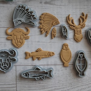 May include: A collection of plastic cookie cutters and matching tan dough cutouts featuring various shapes including a buffalo head, a fish, a deer head, a bird, and a feathered headdress. The set includes both the gray plastic molds and the shapes.