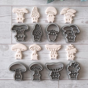 May include: An assortment of mushroom-shaped cookie cutters and cookies. The cutters are gray, and the cookies are off-white. The image is on a light wood-grain surface. The cutters and cookies are arranged in rows.