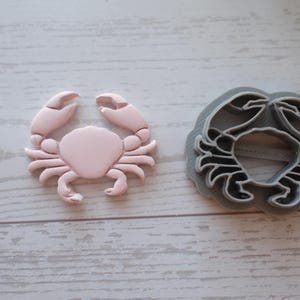 Puede incluir: Un cortador de galletas con forma de cangrejo rosa claro y un cortador de plástico gris. El cangrejo es de color rosa claro con pinzas y un cuerpo redondeado. El cortador gris tiene forma de cangrejo, con un contorno detallado.