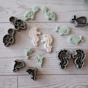 Könnte beinhalten: Eine Auswahl an Ausstechern in Form von Meerestieren. Die Sammlung umfasst Schildkröten, Seepferdchen, Delfine und Wale in verschiedenen Farben, darunter Grau, Weiß und Mintgrün. Die Ausstecher sind auf einer hellen Holzmaserung angeordnet.
