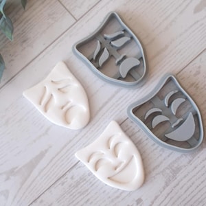 May include: Four cookie cutters in the shape of theater masks, two in white and two in gray. The white masks are the baked cookies, and the gray masks are the cutters. The masks are arranged on a light wood-grain surface.