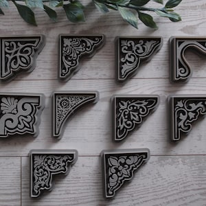 May include: A collection of ten gray and black corner brackets with ornate, symmetrical designs. The brackets are arranged on a light wood-grain surface, showcasing their decorative potential for home decor or craft projects.