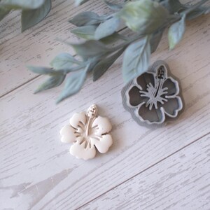 May include: A white hibiscus flower and a gray hibiscus flower-shaped cookie cutter are displayed on a white wooden surface. The flower has detailed petals and a central stamen. A sprig of green leaves is in the background.