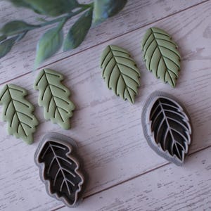 Puede incluir: Una vista aérea muestra varias piezas de arcilla polimérica con forma de hoja verde claro y dos cortadores con forma de hoja. Las hojas tienen patrones de venas detallados. Los cortadores son grises con un interior blanco. Unas pocas hojas verdes están en el fondo.