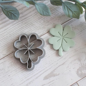 Può includere: Un tagliabiscotti a forma di quadrifoglio in metallo grigio accanto a un quadrifoglio verde chiaro. Il quadrifoglio è su una superficie di legno chiaro con rametti di foglie verdi sullo sfondo. Tema del giorno di San Patrizio.