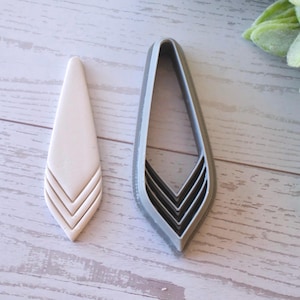 May include: A cream-colored earring and a gray earring cutter sit on a light wood surface. The earring has a teardrop shape with three chevron lines at the bottom. The cutter is the same shape, with a blade to cut the earring.