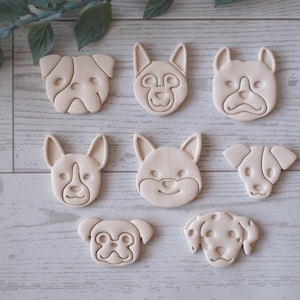 Pode incluir: Uma coleção de cortadores de biscoitos bege em forma de rosto de cachorro. Os cortadores apresentam várias raças de cães, incluindo um buldogue, um pastor alemão e um dálmata. Os cortadores estão dispostos em uma superfície clara com grão de madeira.