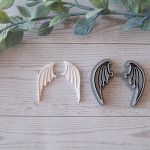 Pode incluir: Dois pares de asas decorativas. Um par é branco, o outro cinza com detalhes pretos. As asas estão dispostas sobre uma superfície clara com grão de madeira, com um raminho de vegetação no fundo. Ideais para artesanato ou bijuteria.