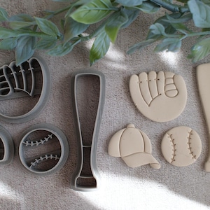 May include: A collection of baseball-themed cookie cutters. The set includes a baseball glove, baseball bat, baseball, baseball cap, and a baseball. The cutters are gray plastic and beige. Green leaves are in the background.