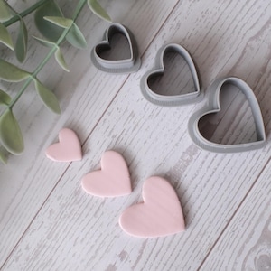 May include: An overhead shot shows heart-shaped cookie cutters and pink heart-shaped decorations on a light wood surface. Green leafy sprigs are in the upper left corner. The cutters are gray, and the hearts are a soft pink.